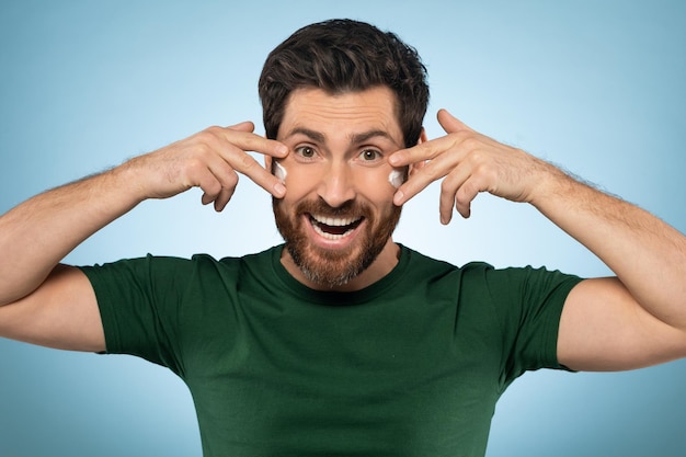 Excited handsome man applying moisturising cream on face standing over blue background doing morning routine