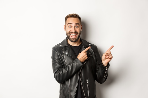 Excited, handsome man in black leather jacket, pointing fingers right at promo offer, showing logo, white background.