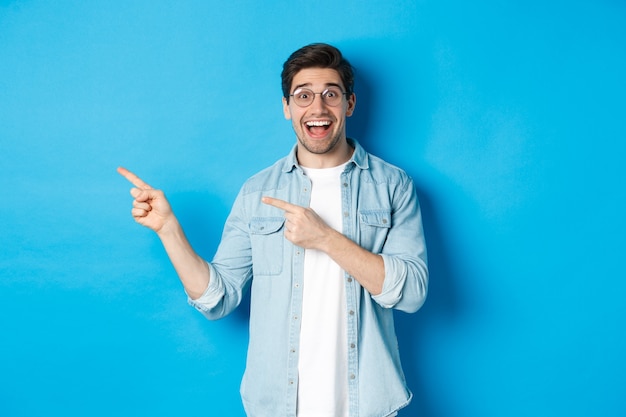 Excited handsome man in glasses pointing fingers left, looking happy while showing advertisement, demonstrating on blue wall