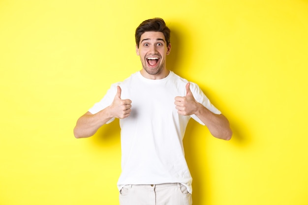 Excited handsome man showing thumbs up, approve and saying yes, standing over yellow wall