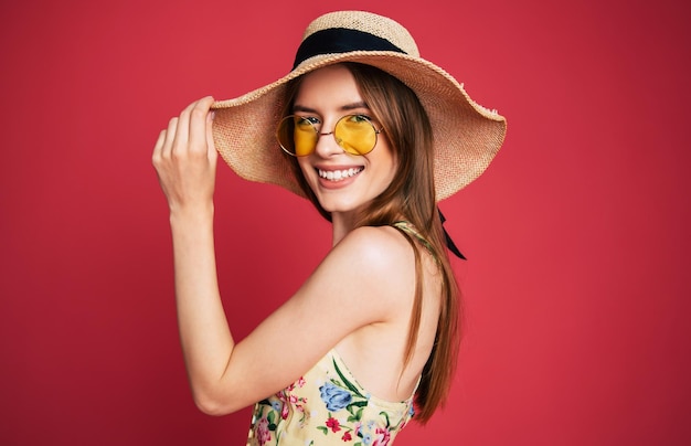 Excited and happy beautiful lovely young woman in sunglasses, dress and summer hat is have a fun while posing on pink background