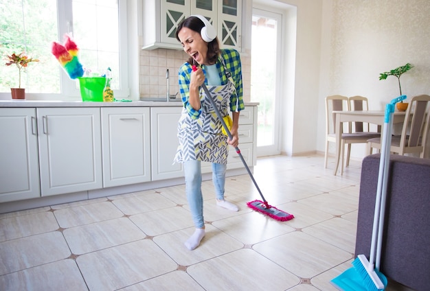 Excited and happy beautiful young woman in an apron is cleaning the floor in her kitchen at home and dancing