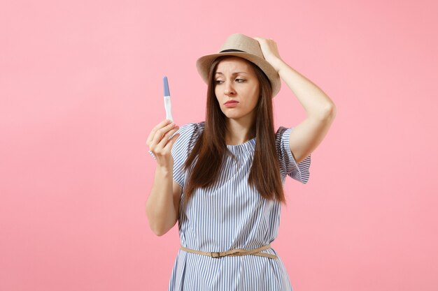 Excited happy woman in blue dress, hat hold in hand, looking at pregnancy test isolated on pink background. Medical healthcare gynecological, pregnancy fertility maternity people concept. Copy space