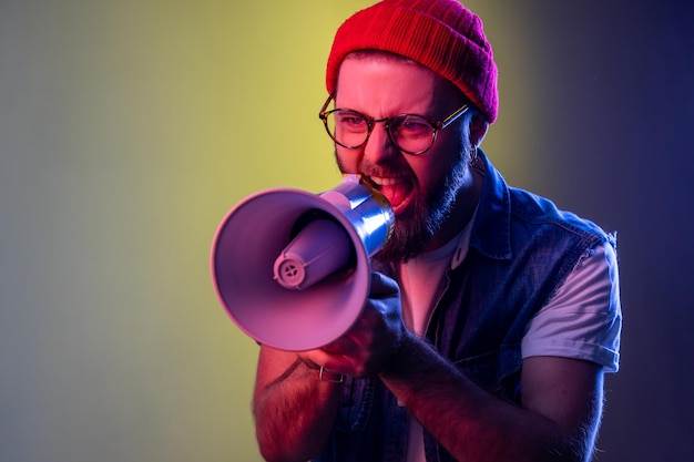 Excited hipster guy screaming loud using megaphone, making announcement, presentation, protesting, wearing beanie hat and denim vest. Indoor studio shot isolated on colorful neon light background.