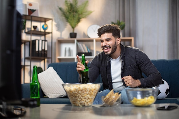 Excited indian man with chips and beer watching soccer on TV