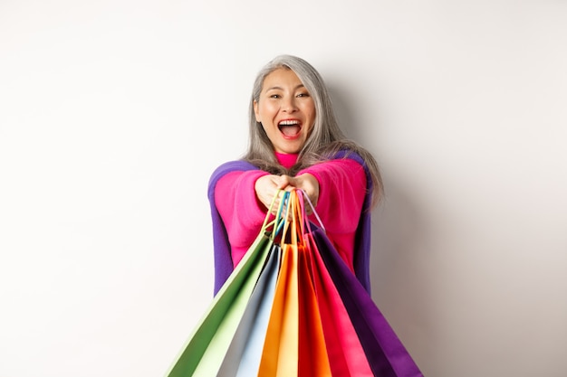 Excited korean lady with grey hair, laughing and showing shopping bags, visit stores with special discounts, standing over white background.