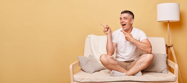 Excited laughing brunette young man wearing casual white t shirt sitting on sofa against beige wall pointing at advertisement area copy space for promotional text