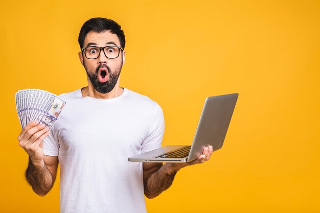 Excited man in casual holding lots of money in dollar currencys and laptop in hands isolated over yellow background.