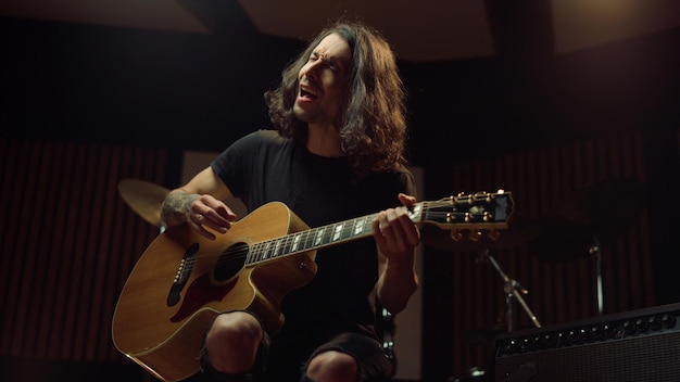 Excited man playing guitar in music studio Cheerful guy singing in dark hall