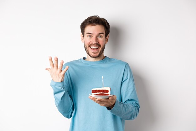 Excited man receive birthday surprise, holding bday cake and smiling happy, standing over white background, making wish on lit candle.