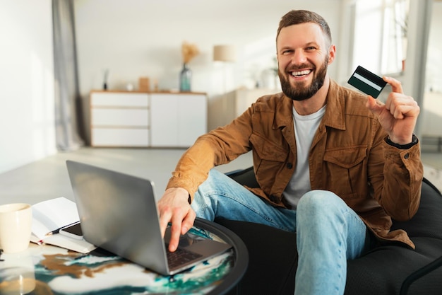 Excited man shopping online showing credit card using laptop indoors