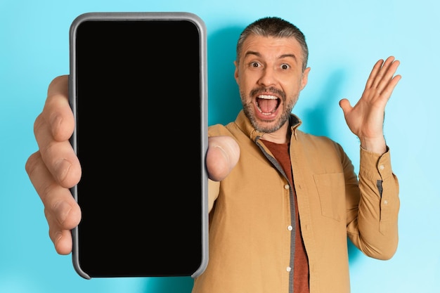 Excited man showing blank phone screen shouting over blue background