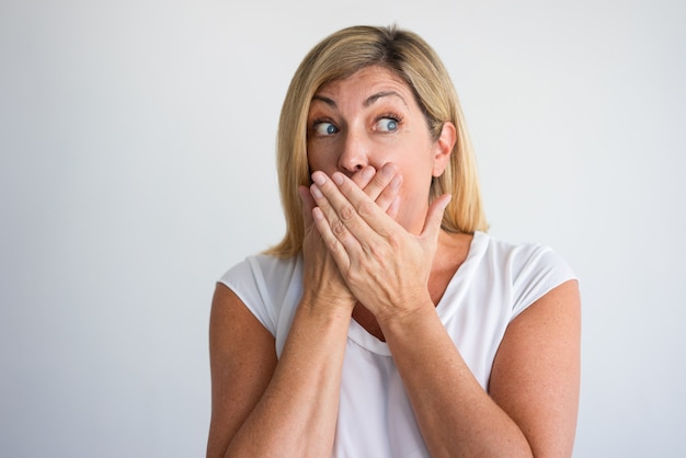 Excited middle aged Caucasian woman covering mouth with both hands at camera.