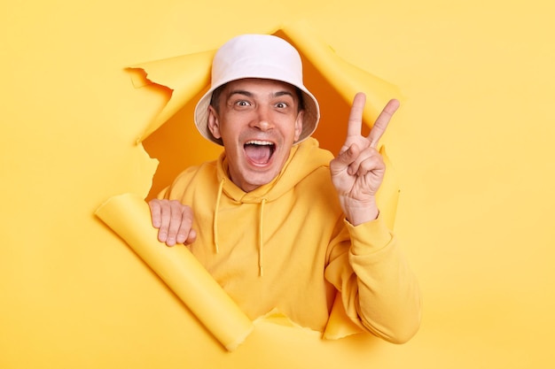 Excited optimistic man wearing casual hoodie and panama looking through torn hole in yellow paper showing v sign victory gesture with fingers looking at camera with toothy smile