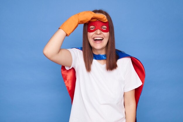 Photo excited overjoyed happy woman wearing white tshirt rubber gloves cloak and eye mask jeeps hand near eyes looking far with laughing face standing isolated over blue background