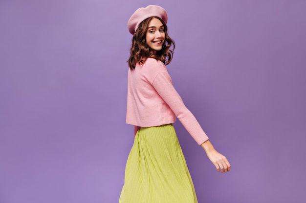 Photo excited parisian woman moves on purple background cheerful curly girl in beret pink sweater and green skirt dances