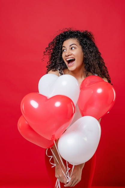 Excited playful black girl with helium balloons isolated on red