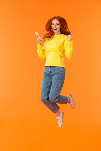 Excited and pleased good-looking ginger woman in sweater, jeans, jumping over orange