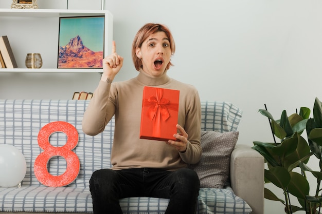 Excited points at up handsome guy on happy women day holding present sitting on sofa in living room