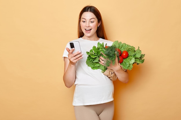 Excited pregnant woman holding fresh vegetables posing isolated over beige background using mobile phone for ordering organic food online