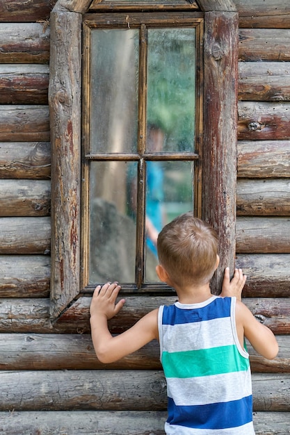 Excited preschooler boy looks into window of ancient log house