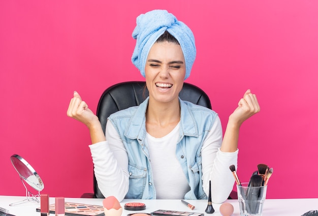 Excited pretty caucasian woman with wrapped hair in towel sitting at table with makeup tools keeping fists up 