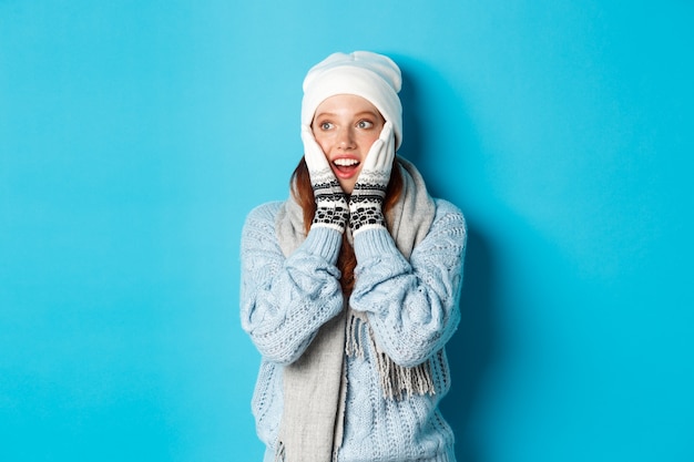 Excited redhead girl staring left at logo, wearing winter clothes, beanie, gloves and sweater, standing over blue background