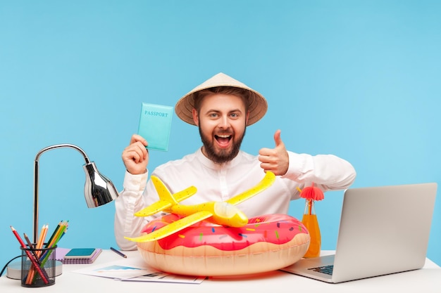 Excited satisfied man office worker sitting at workplace with toy plane and lifebuoy, showing thumbs up holding passport, returned from vacation abroad. Indoor studio shot isolated on blue background