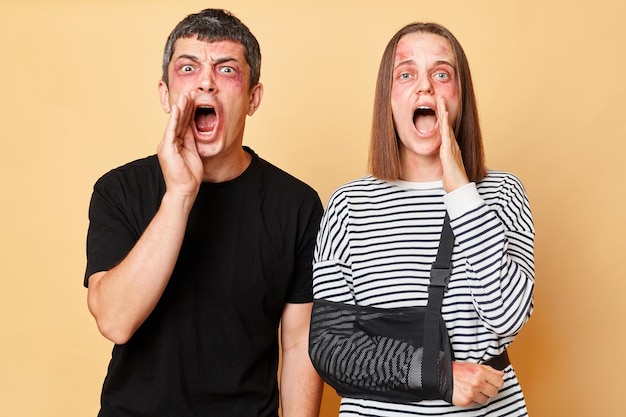 Excited scared shocked couple screaming loud about violence bandit attack injured man and woman with bruise isolated over beige background