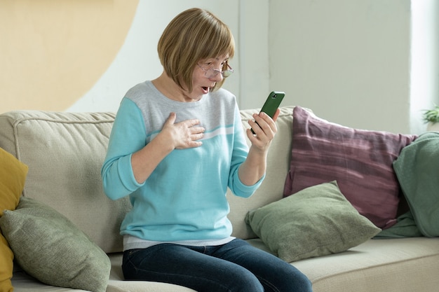 Excited senior woman surprised by good unbelievable news looking at smartphone screen sitting on sofa at home