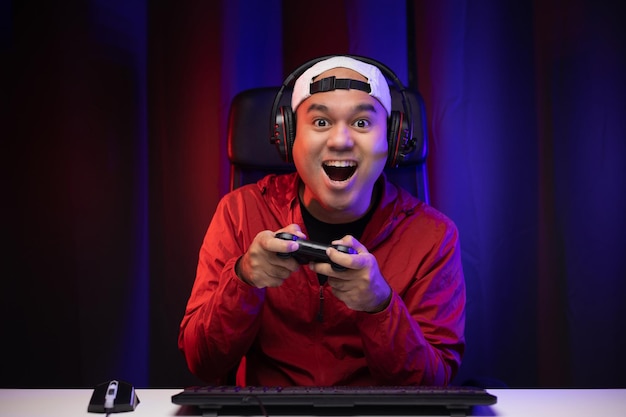 Excited and shocked face of Asian gamer with headphone holding joystick playing video game online sitting on chair at living room. Indian professional gamer streaming on social playing game very fun