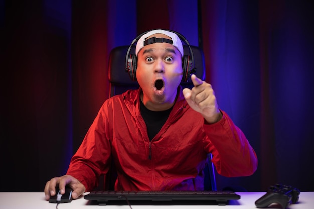 Excited and shocked face of Asian gamer with headphone playing computer pc video game online sitting at living room. Indian professional gamer streaming on social playing game very fun