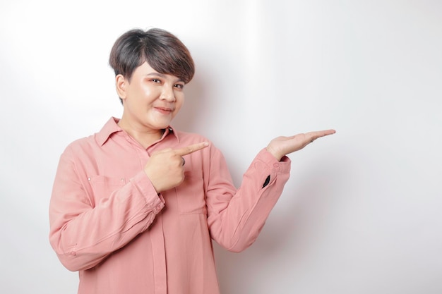 Excited shorthaired Asian woman wearing a pink shirt pointing at the copy space beside her isolated by a white background