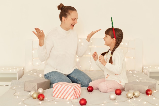 Excited smiling happy mother and daughter wearing casual style white sweaters sitting on bed with Christmas decorations, yelling happily, celebrating new year.