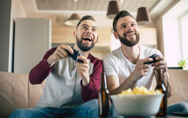 Excited smiling men playing in video games on tv at home on the couch. Friends with joysticks play game with happy emotions on faces