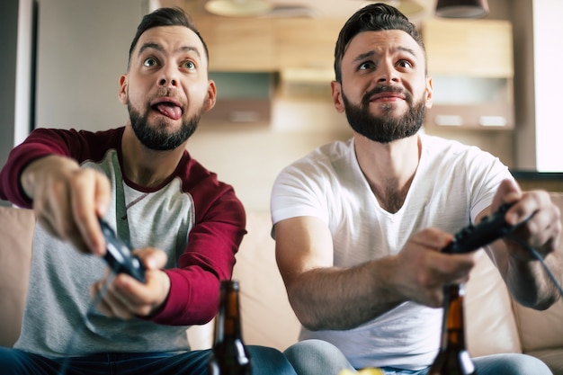 Excited smiling men playing in video games on tv at home on the couch. Friends with joysticks play game with happy emotions on faces