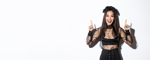 Excited smiling woman celebrating halloween in black gothic dress and wreat Asian girl wearing witch costume and pointing fingers up standing white background