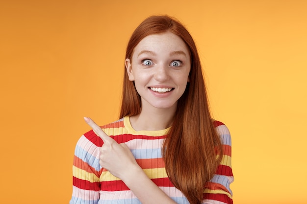 Excited thrilled good-looking young silly redhead girl surprised pop eyes camera gasping impressed pointing upper left corner see super star grinning excitement delight, orange background