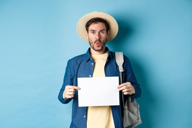 Excited tourist in straw hat, holding empty piece of paper and looking amused, going on summer travel, standing on blue background.