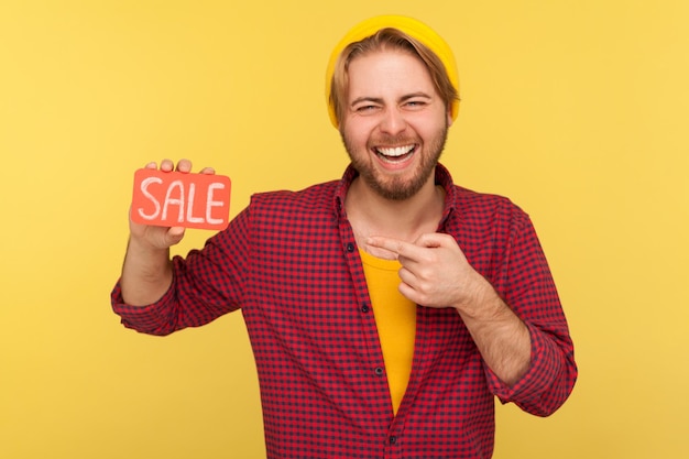 Excited trendy hipster guy in checkered shirt pointing at Sale word and laughing loudly, feeling carefree amused by discounts, Black Friday low prices. indoor studio shot isolated on yellow background