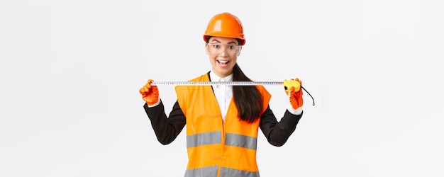 Excited and upbeat asian female construction engineer measuring layout holding tape measure and smiling ready for work at build something standing over white background in safety helmet