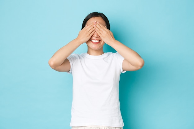 Excited and upbeat asian girl in white t-shirt making wish, close eyes as awaiting for surprise or playing hide-n-seek, standing upbeat over blue wall, blindfolded with happy smile