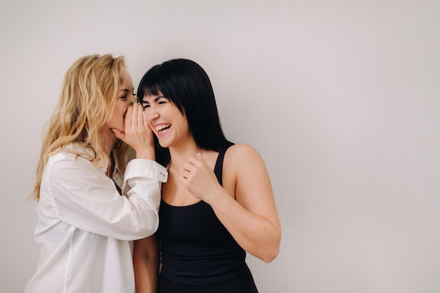 An excited woman in a black swimsuit listens to a friend in a shirt whispering in her ear on a light background