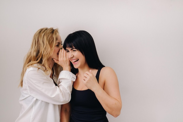 An excited woman in a black swimsuit listens to a friend in a shirt whispering in her ear on a light background