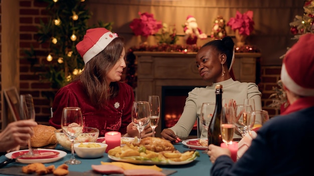 Excited woman discussing with close friend at Christmas dinner while drinking champagne. Festive diverse people celebrating winter feast with sparkling wine and traditional home cooked food.