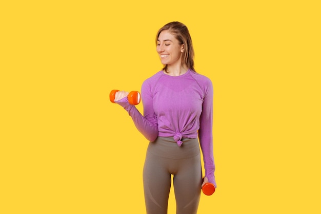 Excited woman is exercising arms with dumbbells over yellow background