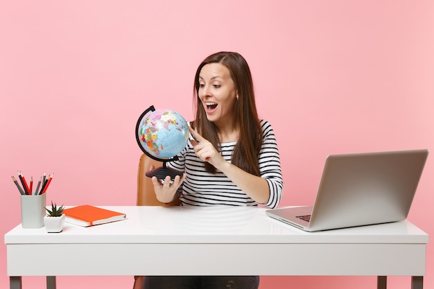 Excited woman looking on world globe planning vacation while sit and work at white desk with contemporary pc laptop