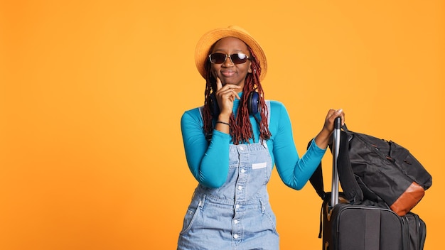 Excited woman wearing sunglasses and hat, feeling confident travelling to holiday destination. Young adult with luggage and trolley bags leaving on urban adventure, cool glasses.