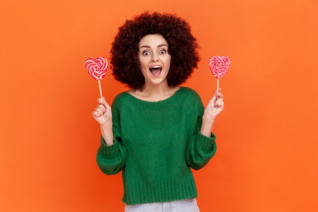 Photo excited woman with afro hairstyle in green casual style sweater holding two candies with heart shape looking at camera with happy positive expression indoor studio shot isolated on orange background