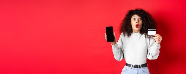 Photo excited woman with credit card showing empty smartphone screen gasping amazed standing against red b